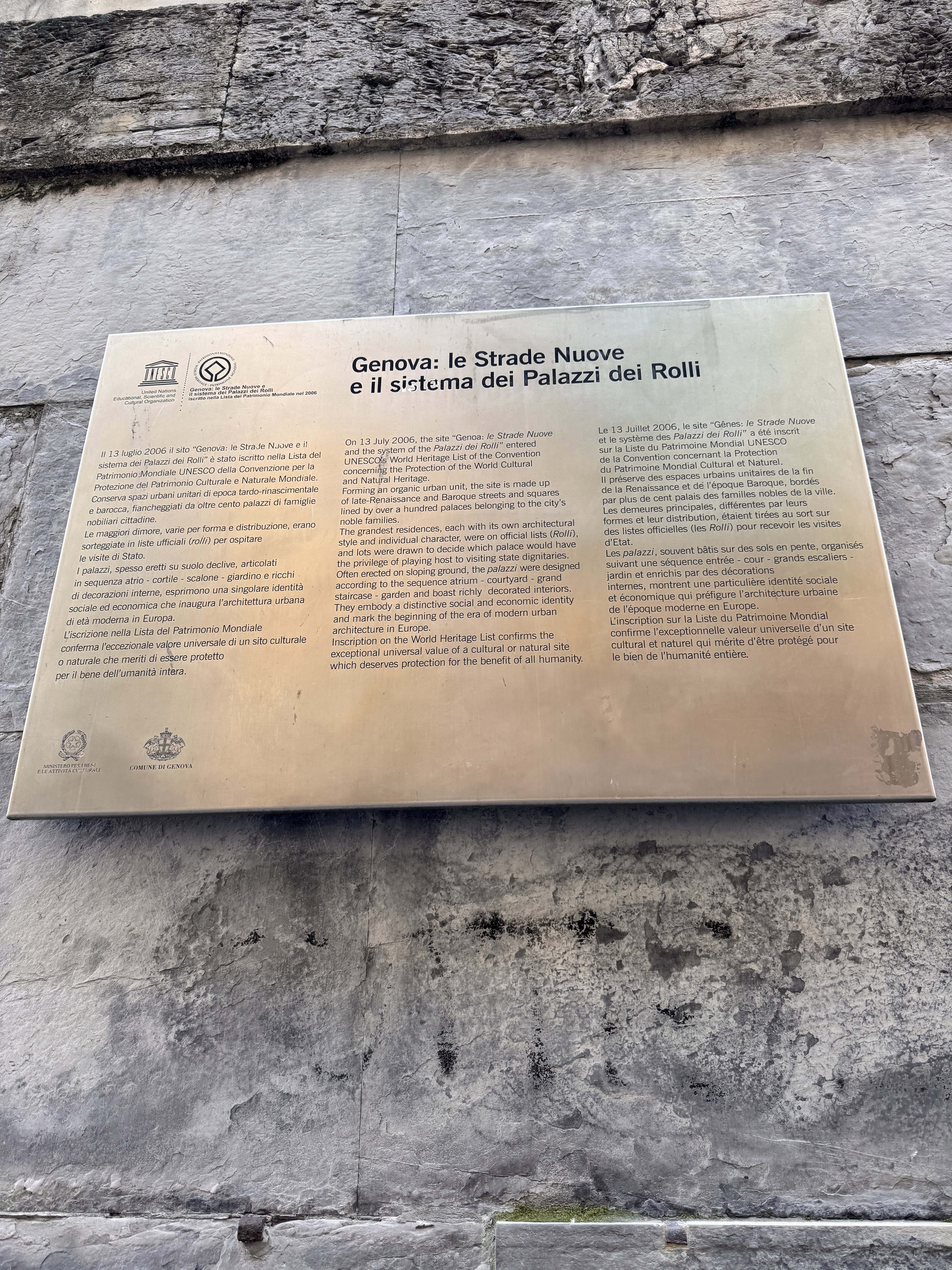 A metal plaque on a stone wall in Genoa, Italy, describes Genova’s Strade Nuove and the Palazzi dei Rolli system in three languages, perfect for visitors exploring the city in 24 hours.