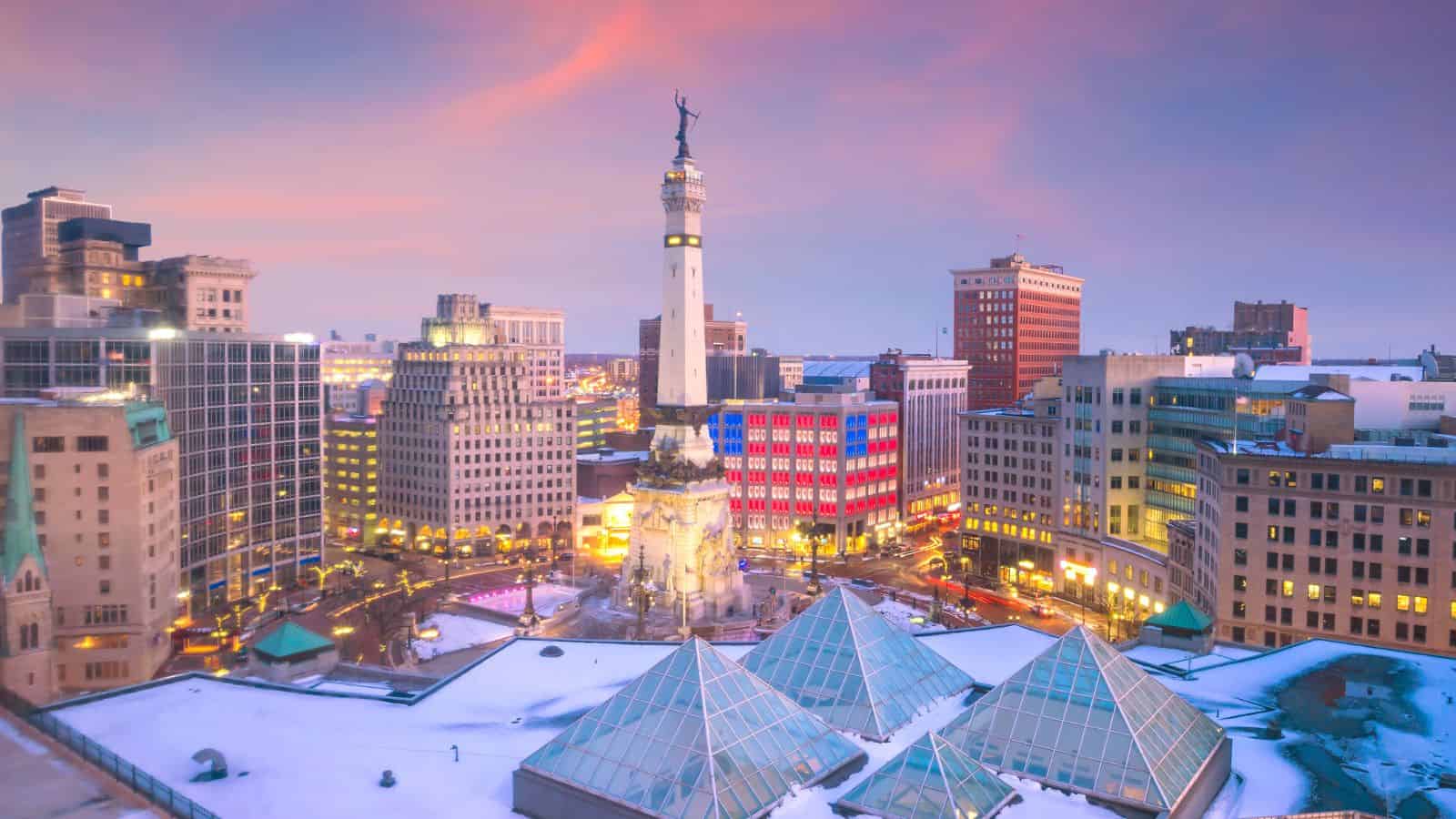 A cityscape at sunset featuring a monument in a snowy downtown surrounded by tall buildings and glowing lights.