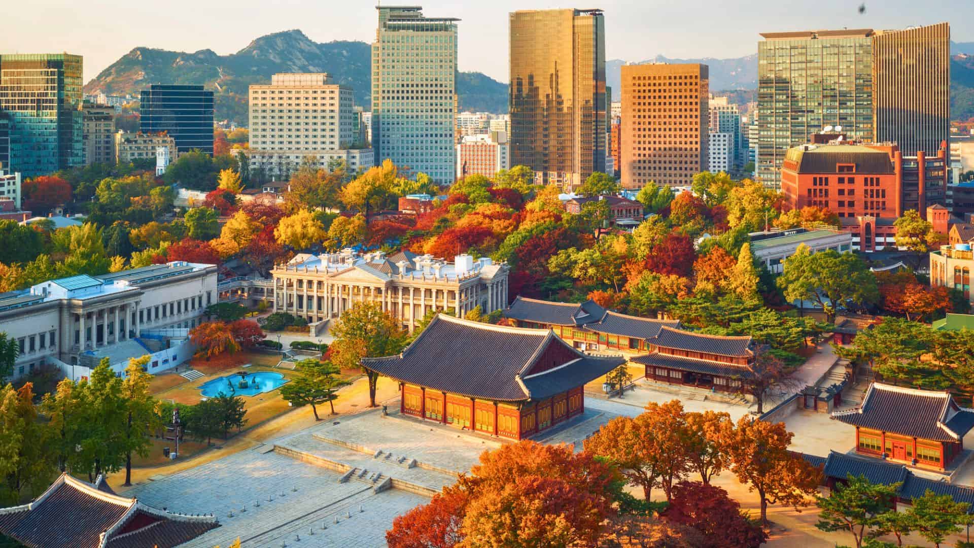 Aerial view of Deoksugung Palace and colorful autumn trees with modern Seoul skyscrapers in the background.