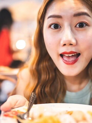 Woman with long hair smiling and holding a plate of food at a lively restaurant.