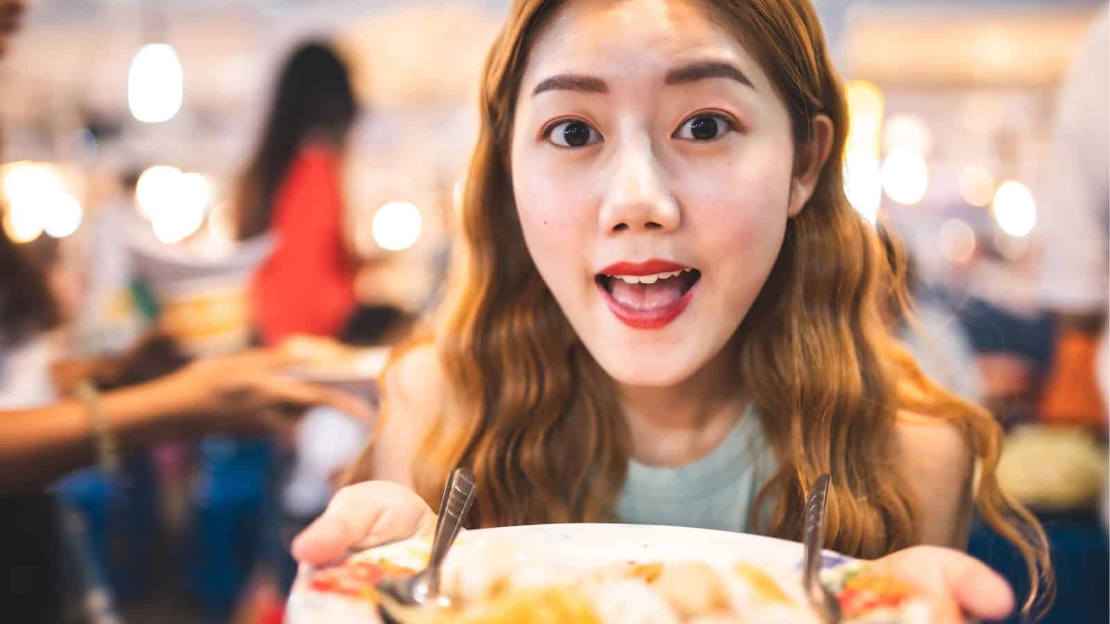 Young woman with long hair smiling and holding a plate of food, looking excited in a busy restaurant.