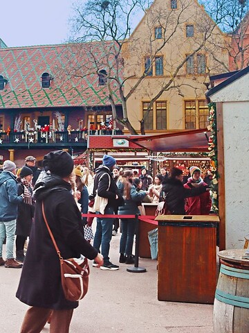 People walk and gather at a festive outdoor market with colorful historic buildings in the background.