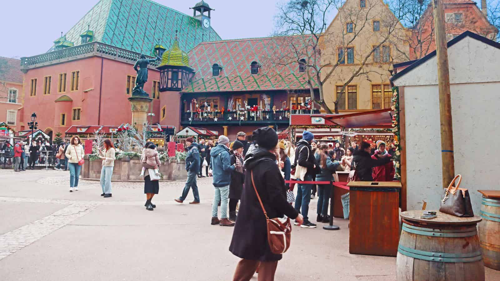 People walk and gather at a festive outdoor market with colorful historic buildings in the background.