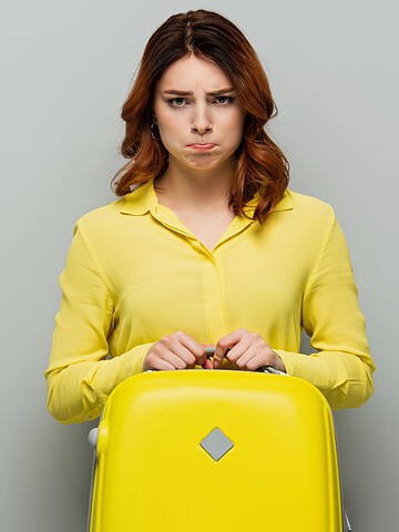 Woman in a yellow shirt holding a yellow suitcase and pouting against a plain gray background.