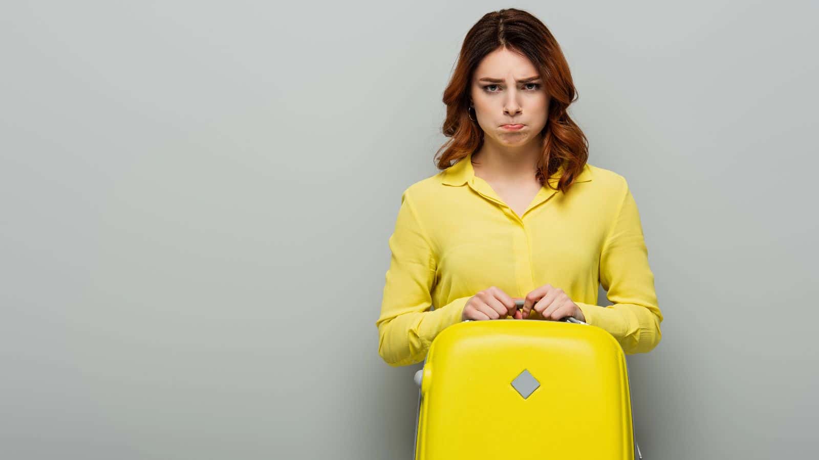 Woman in a yellow shirt holding a yellow suitcase and pouting against a plain gray background.