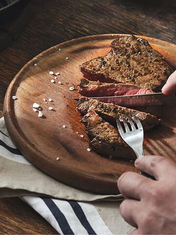 A person cuts a medium-rare steak on a wooden board, with a glass of whiskey and a bowl of salt nearby.