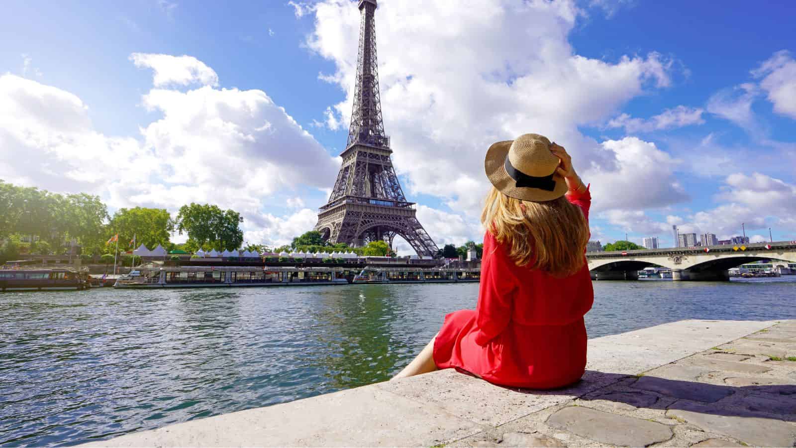 A woman in a red dress and hat sits by the Seine, facing the Eiffel Tower on a sunny day.