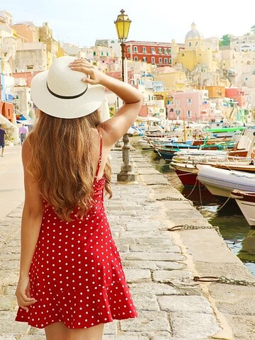 Woman in a red polka-dot dress and hat walks by colorful buildings and boats along a sunny waterfront.