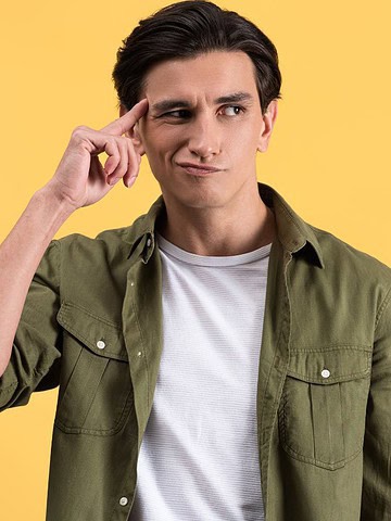 Man in green shirt touching temple and looking up thoughtfully against a yellow background.