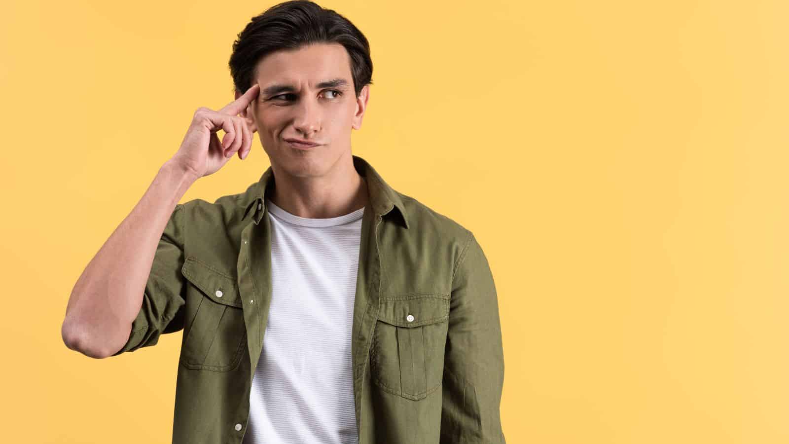 Man in green shirt touching temple and looking up thoughtfully against a yellow background.