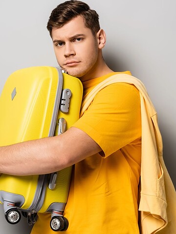 Man in a yellow shirt hugging a yellow suitcase with a neutral expression against a gray background.
