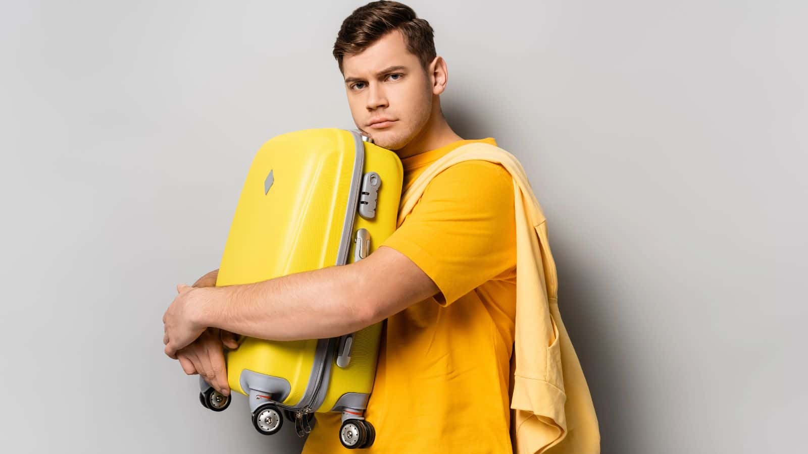 Man in a yellow shirt hugging a yellow suitcase with a neutral expression against a gray background.