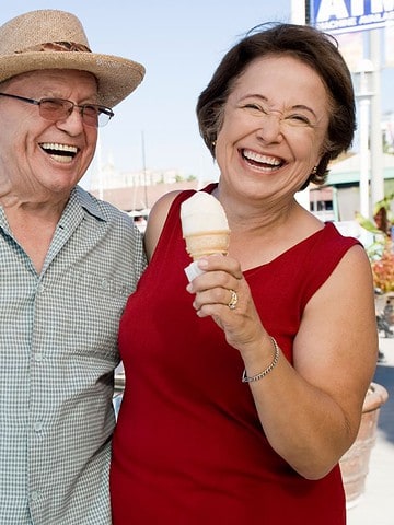 Smiling older couple holding ice cream cones at a marina on a sunny day.