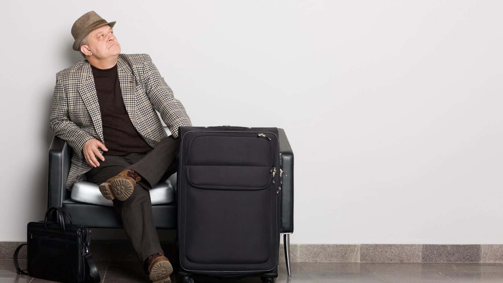 Man in a hat and checked blazer sitting on a black chair with luggage and a briefcase, looking to the side.