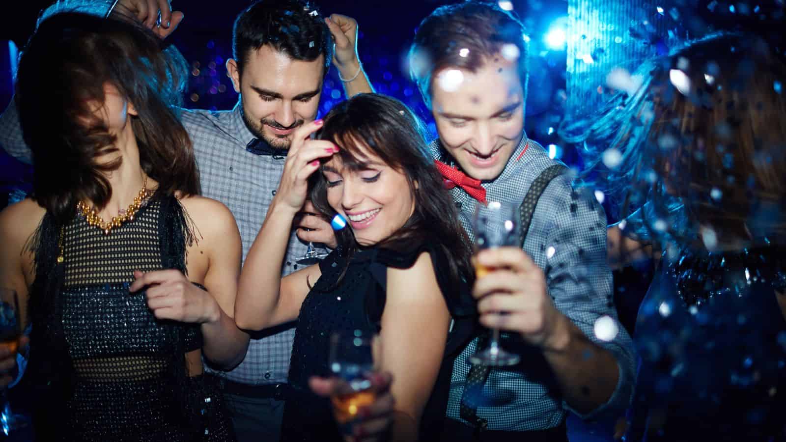 Four people dancing and smiling together at a lively party with drinks and blue lights in the background.