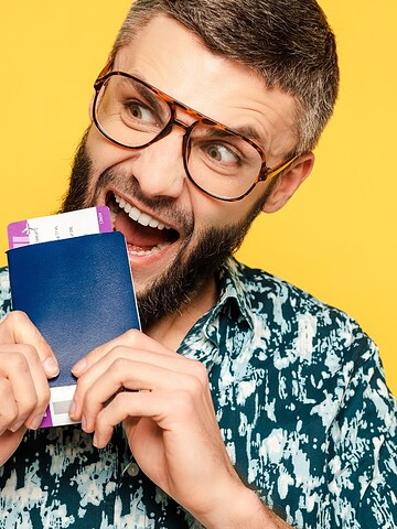 Excited man in glasses playfully biting a passport and boarding pass against a yellow background.