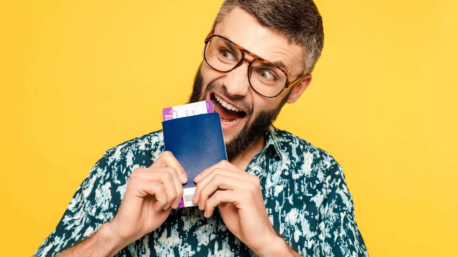 Excited man in glasses playfully biting a passport and boarding pass against a yellow background.