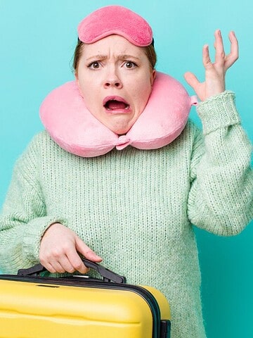 Woman with travel pillow, sleep mask, and suitcase looks confused and frustrated against blue background.