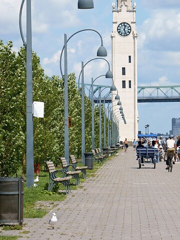 People walk and bike along a riverside path with a clock tower and bridge in the background on a sunny day.