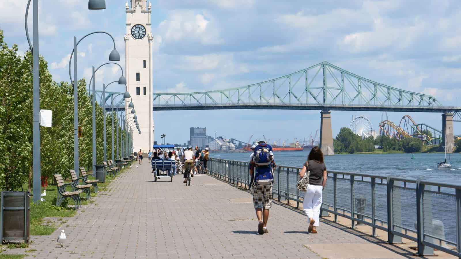 People walk and bike along a riverside path with a clock tower and bridge in the background on a sunny day.