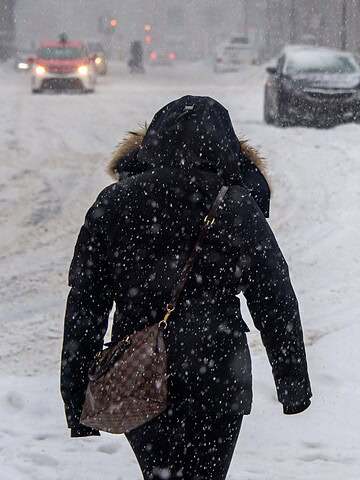 Person in a hooded coat walking on a snowy sidewalk during a snowstorm, cars and people in the background.
