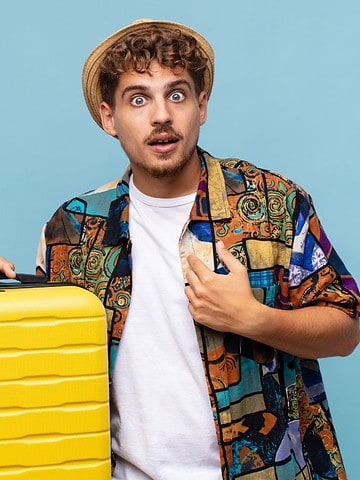 Surprised young man in a hat and colorful shirt holding a yellow suitcase against a blue background.