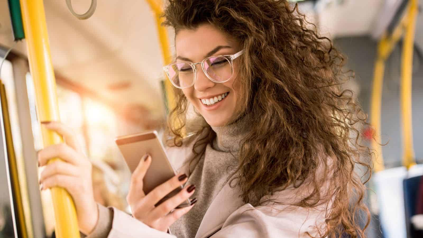 A woman with curly hair smiles while looking at her phone on a bus, holding a yellow handrail.