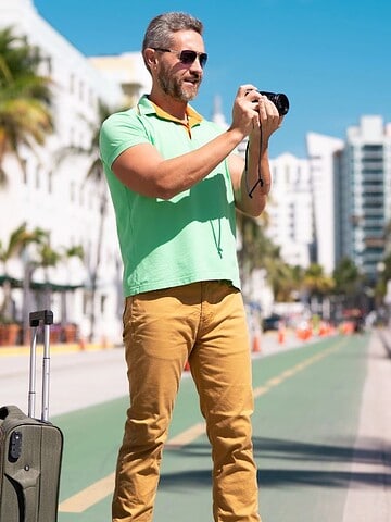 Smiling man with suitcase takes a photo on a sunny city street lined with palm trees and buildings.