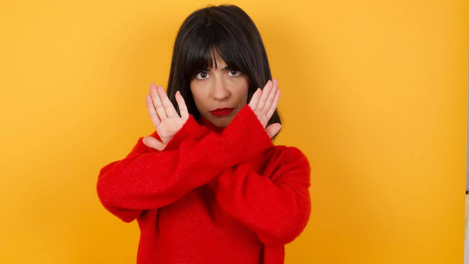 Woman in a red sweater crossing her arms in front of her chest against a yellow background, expressing concern—perhaps thinking about travel safety in December or which countries to avoid during the holiday season.