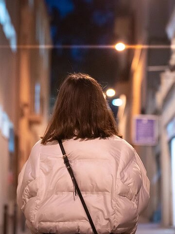 Person with shoulder-length hair in a white jacket walks down a narrow, dimly lit street at night.