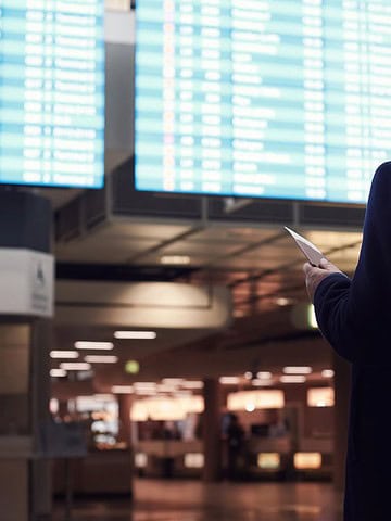 A man in a coat checks his phone while looking at large flight information boards in an airport.