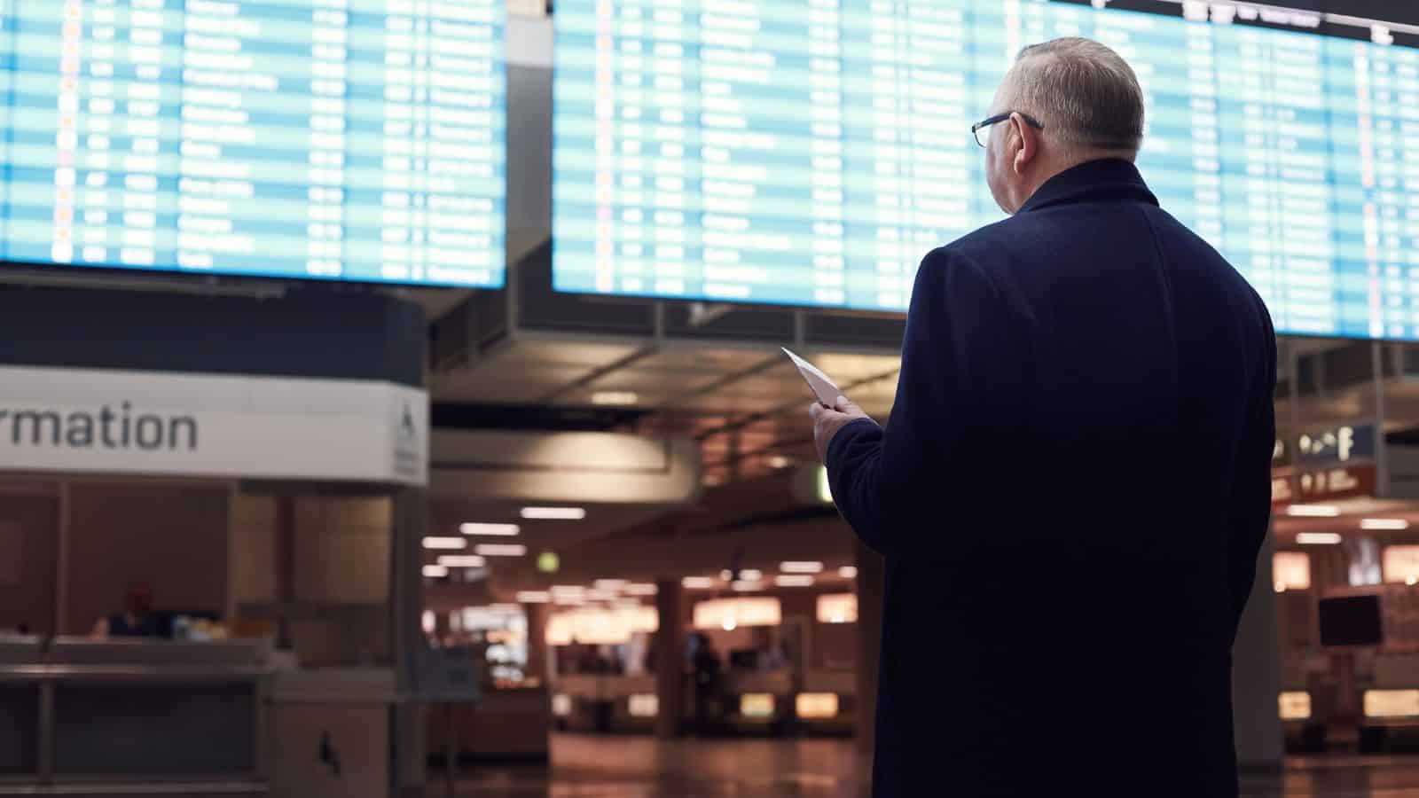 A man in a coat checks his phone while looking at large flight information boards in an airport.