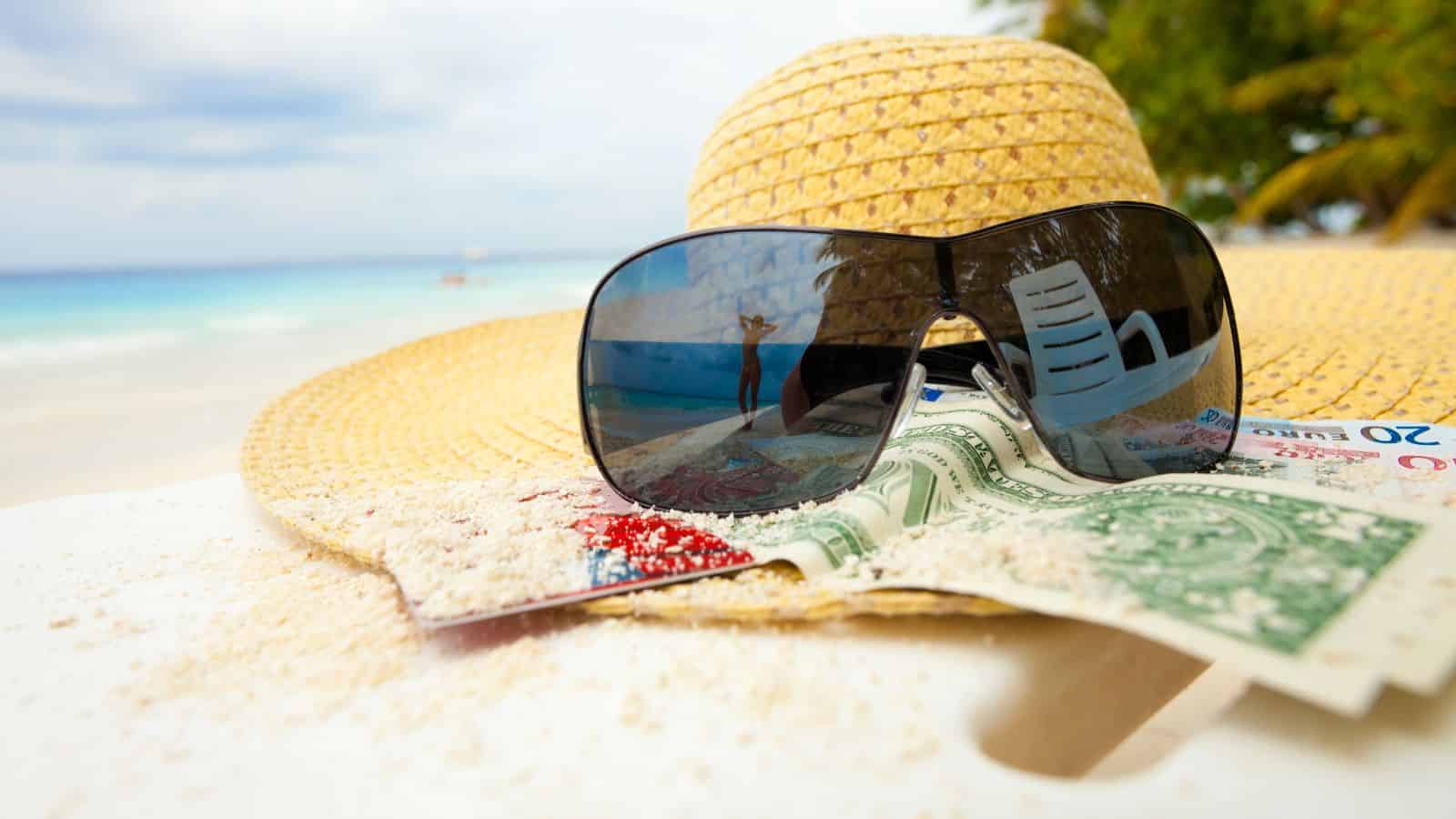 A straw hat, sunglasses, and money on a sandy beach table with the ocean and trees in the background.