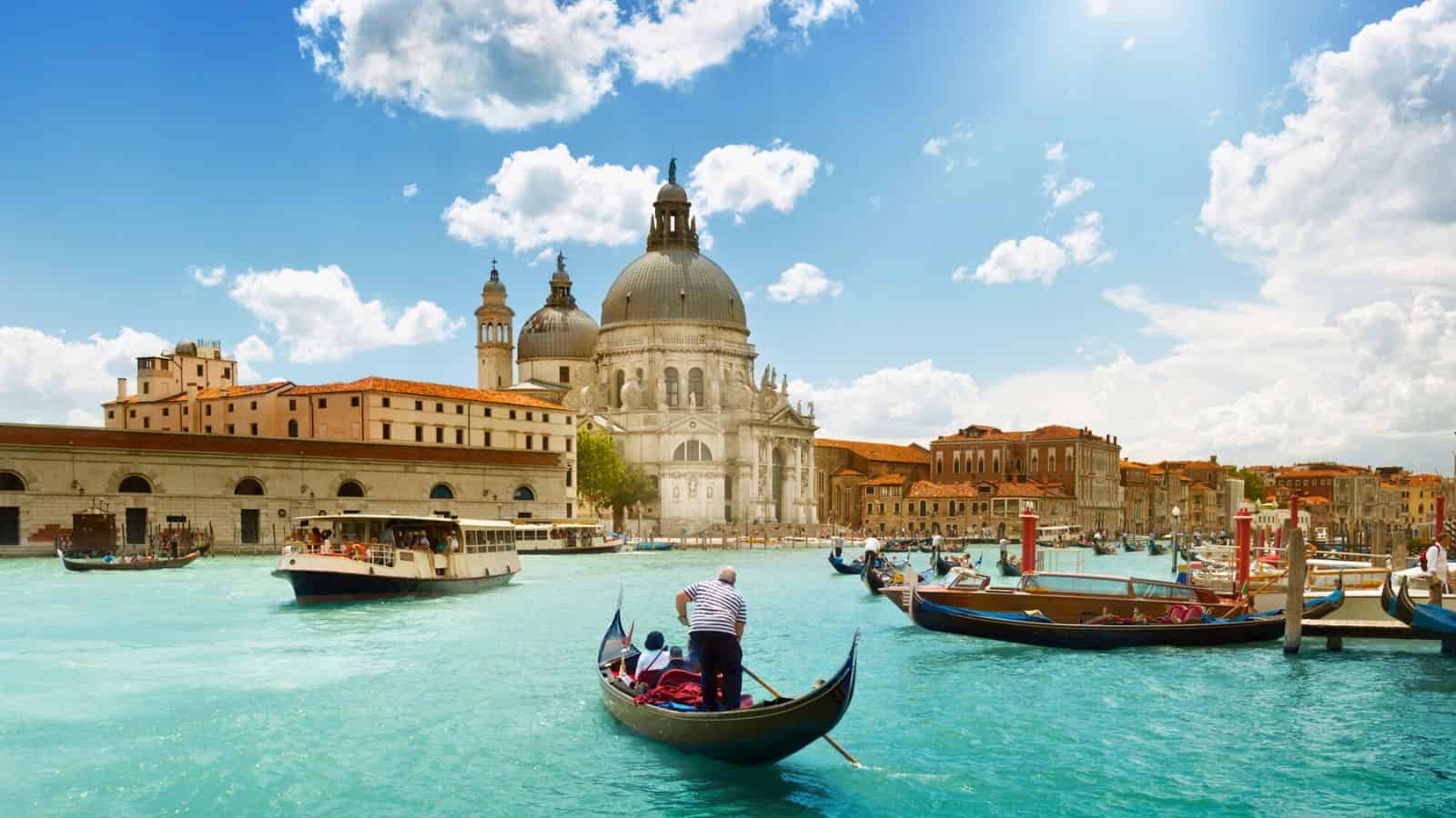 Gondolas and boats on a canal in Venice, Italy, with historic buildings and a domed church under a sunny sky.