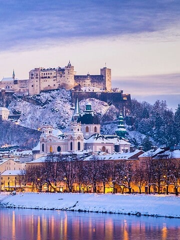 A snowy Salzburg cityscape at dusk with Hohensalzburg Fortress lit up on a hill above the river.