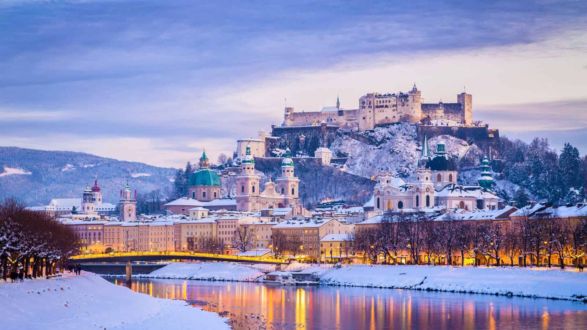 A snowy Salzburg cityscape at dusk with Hohensalzburg Fortress lit up on a hill above the river.