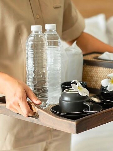 Person holding a tray with water bottles, cups, and flowers in a hotel room with a neatly made bed.