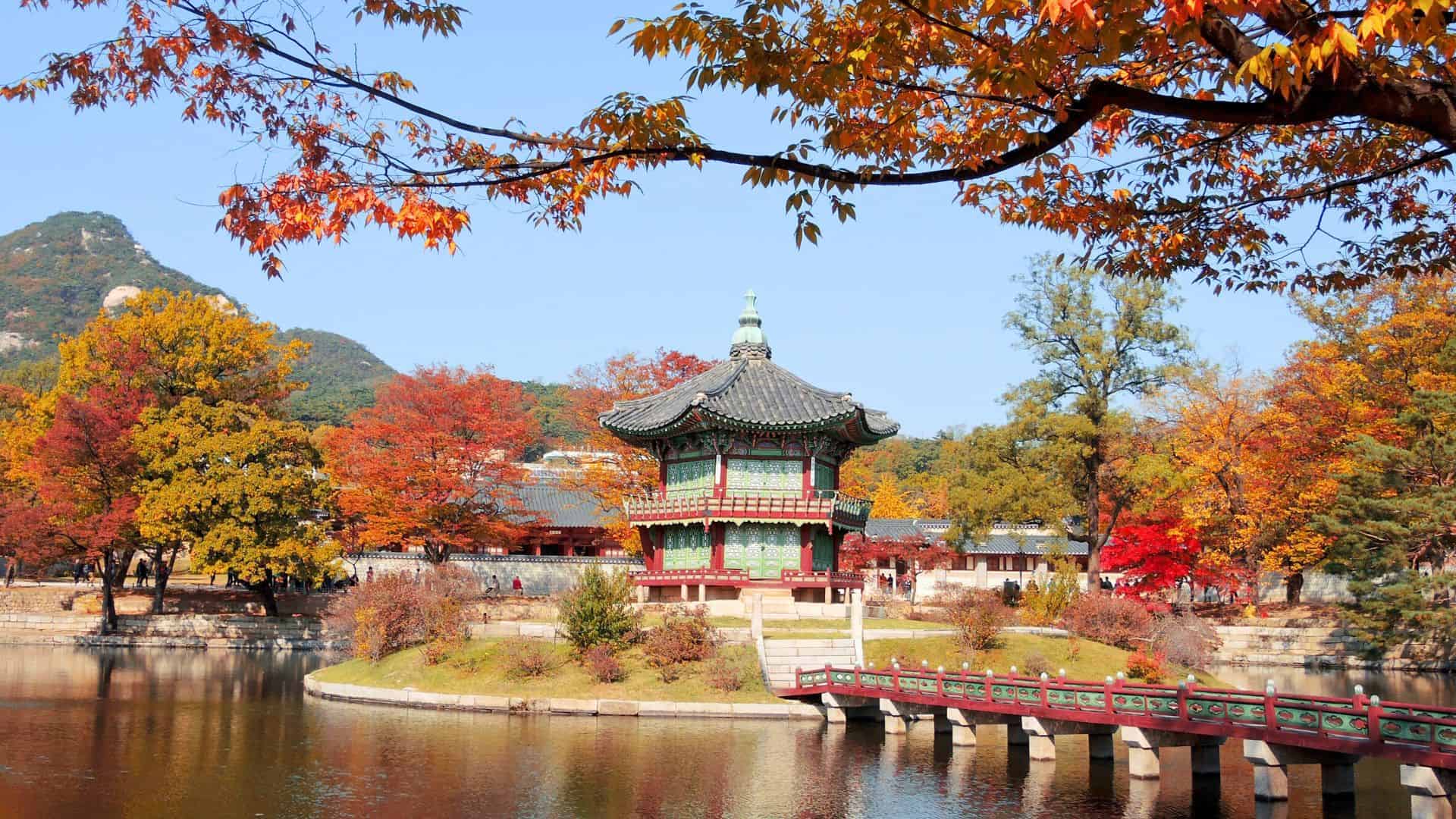 A traditional Korean pavilion by a pond, surrounded by autumn trees with red and orange leaves.
