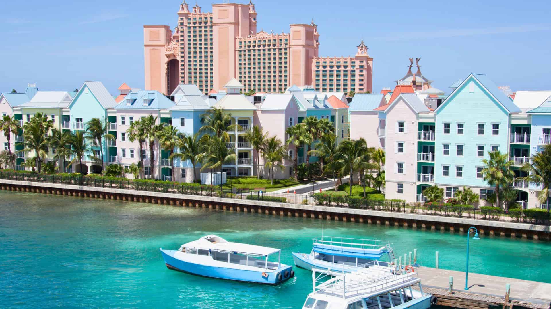 Colorful waterfront buildings and boats with a large pink resort in the background under a clear blue sky.