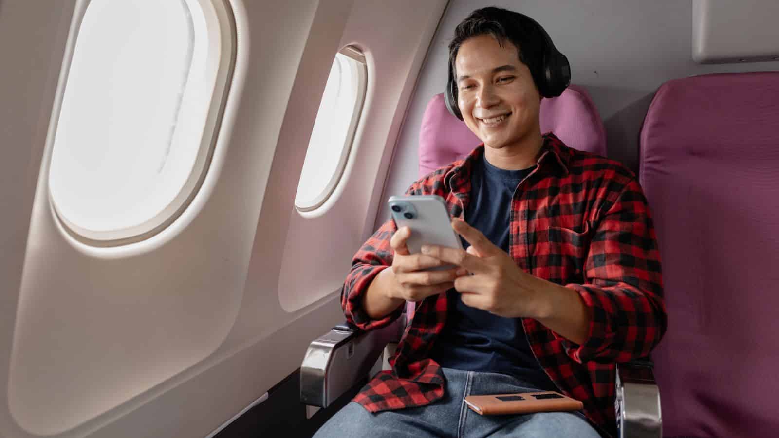 Smiling person with headphones uses a smartphone while sitting on an airplane by the window.