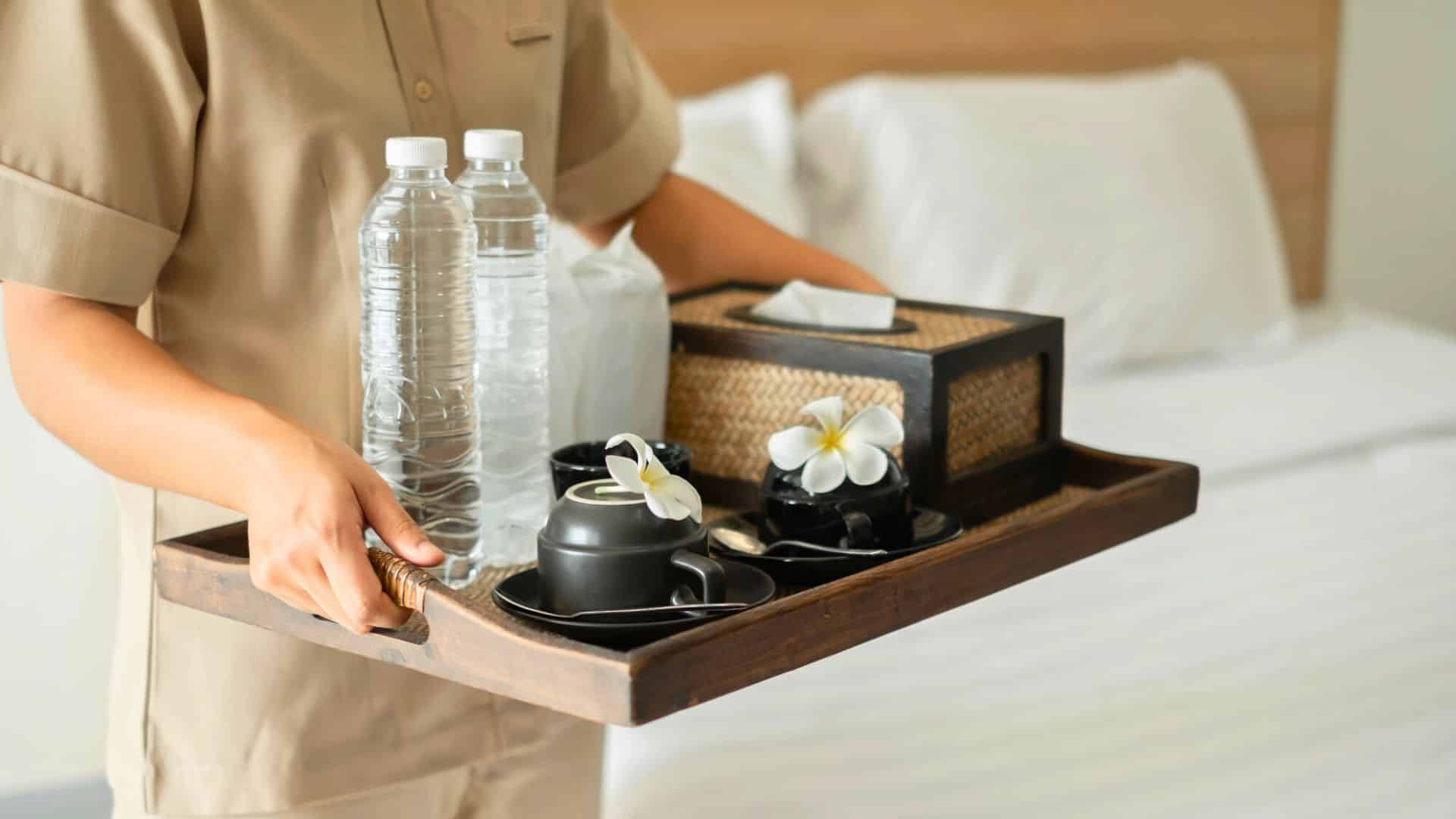 Person holding a tray with water bottles, cups, and flowers in a hotel room with a neatly made bed.