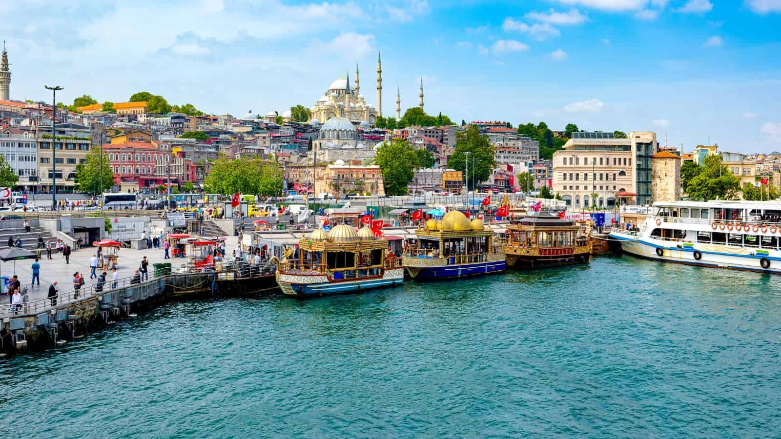 Colorful boats docked at a lively waterfront in Istanbul with mosques and city buildings in the background.
