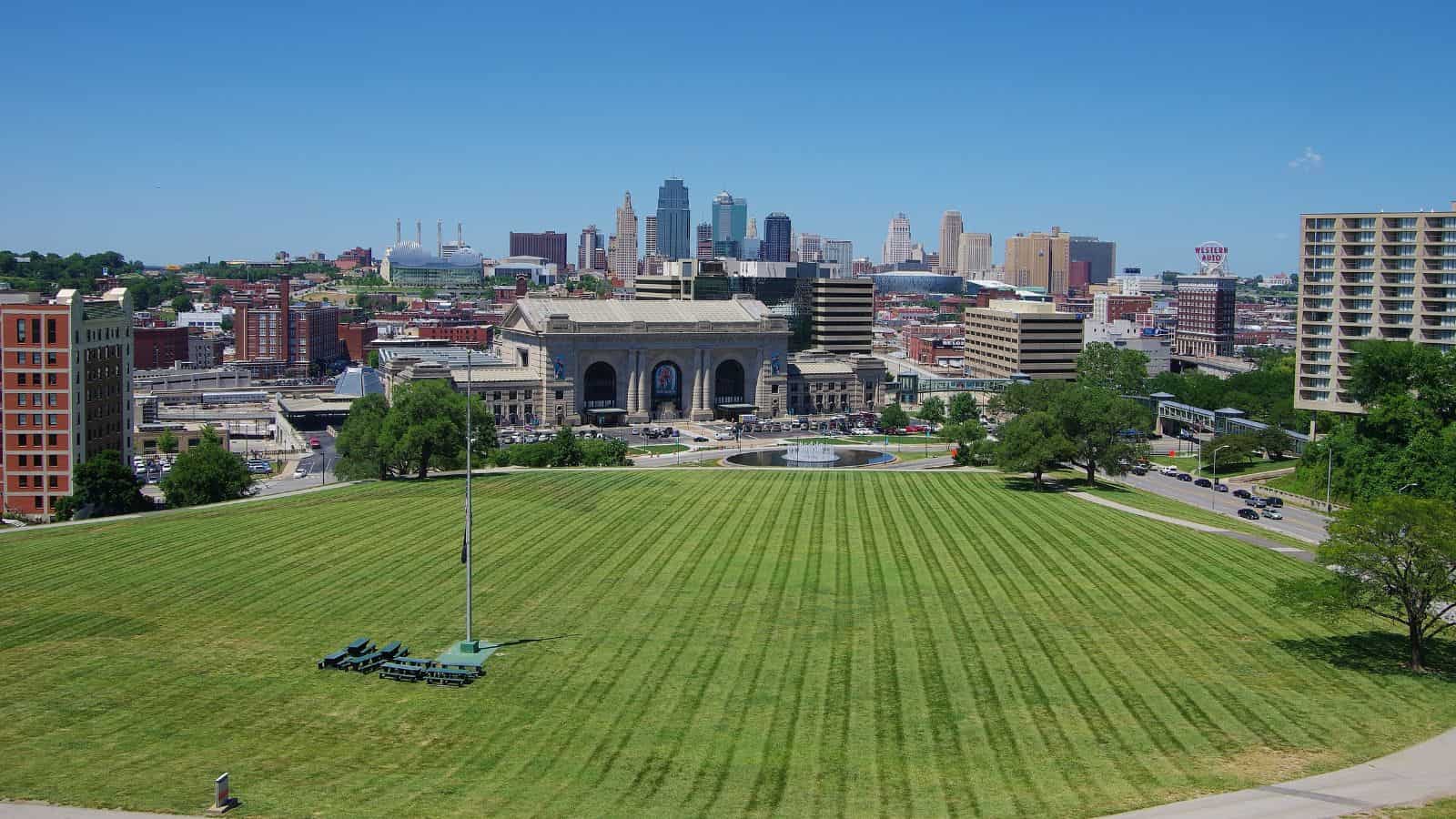 Wide view of a grassy park with a flagpole, city buildings, and blue sky in the background.