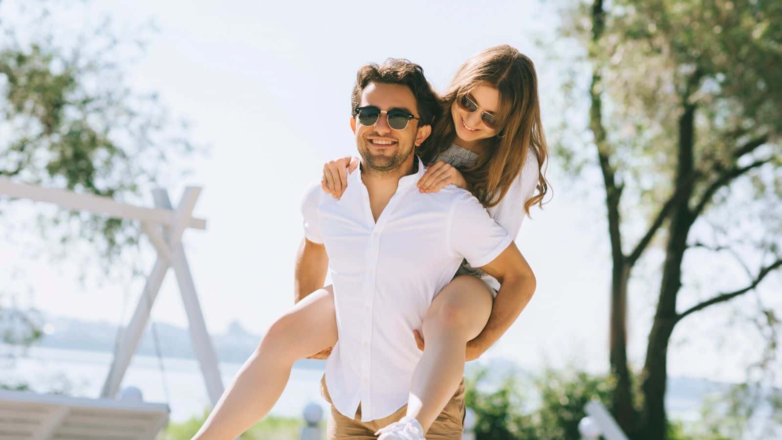 A man gives a woman a piggyback ride outdoors on a sunny day; both are smiling and wearing sunglasses.