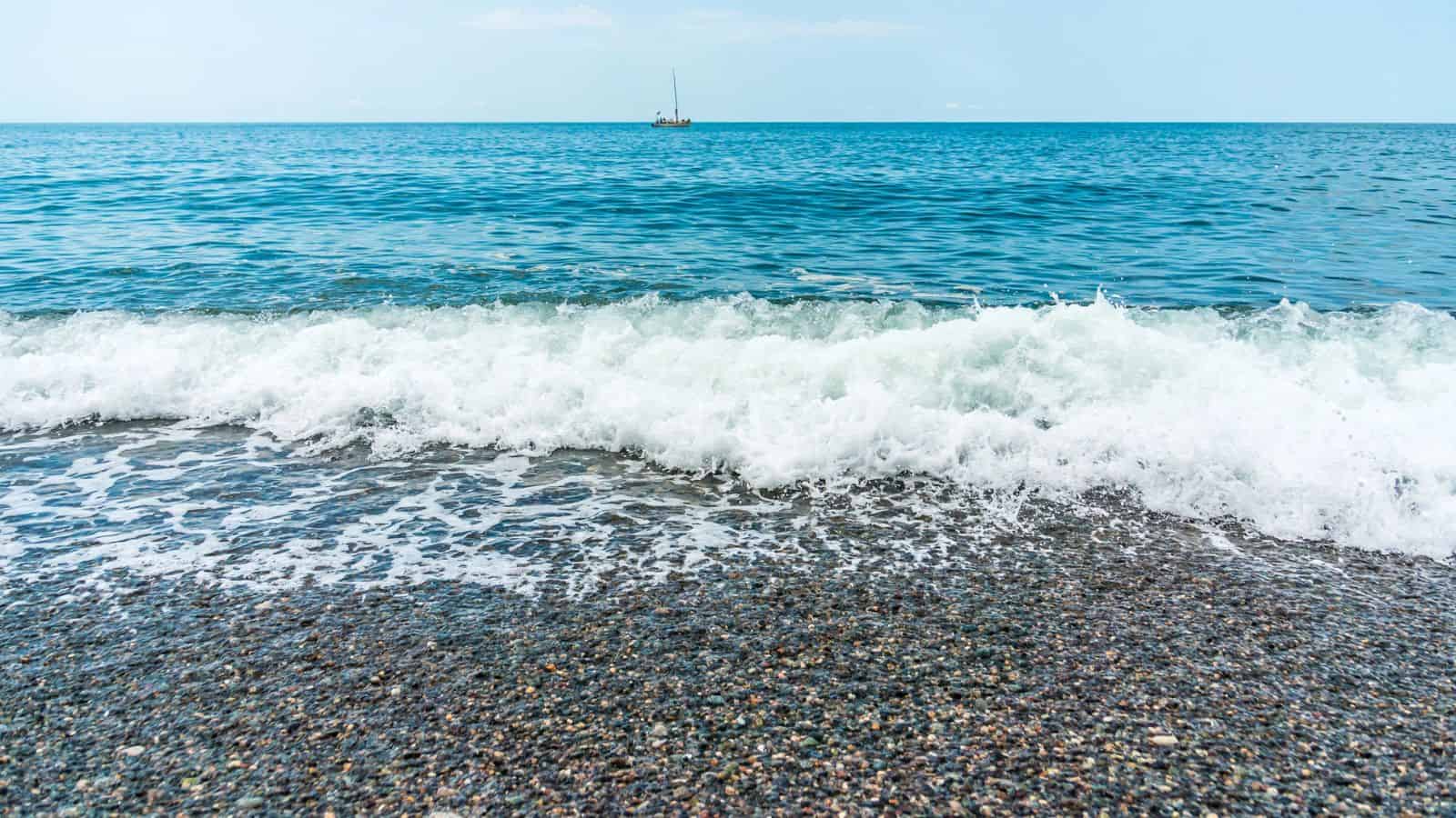 Waves crashing onto a pebbled shore with a sailboat in the distance on a calm, blue sea.