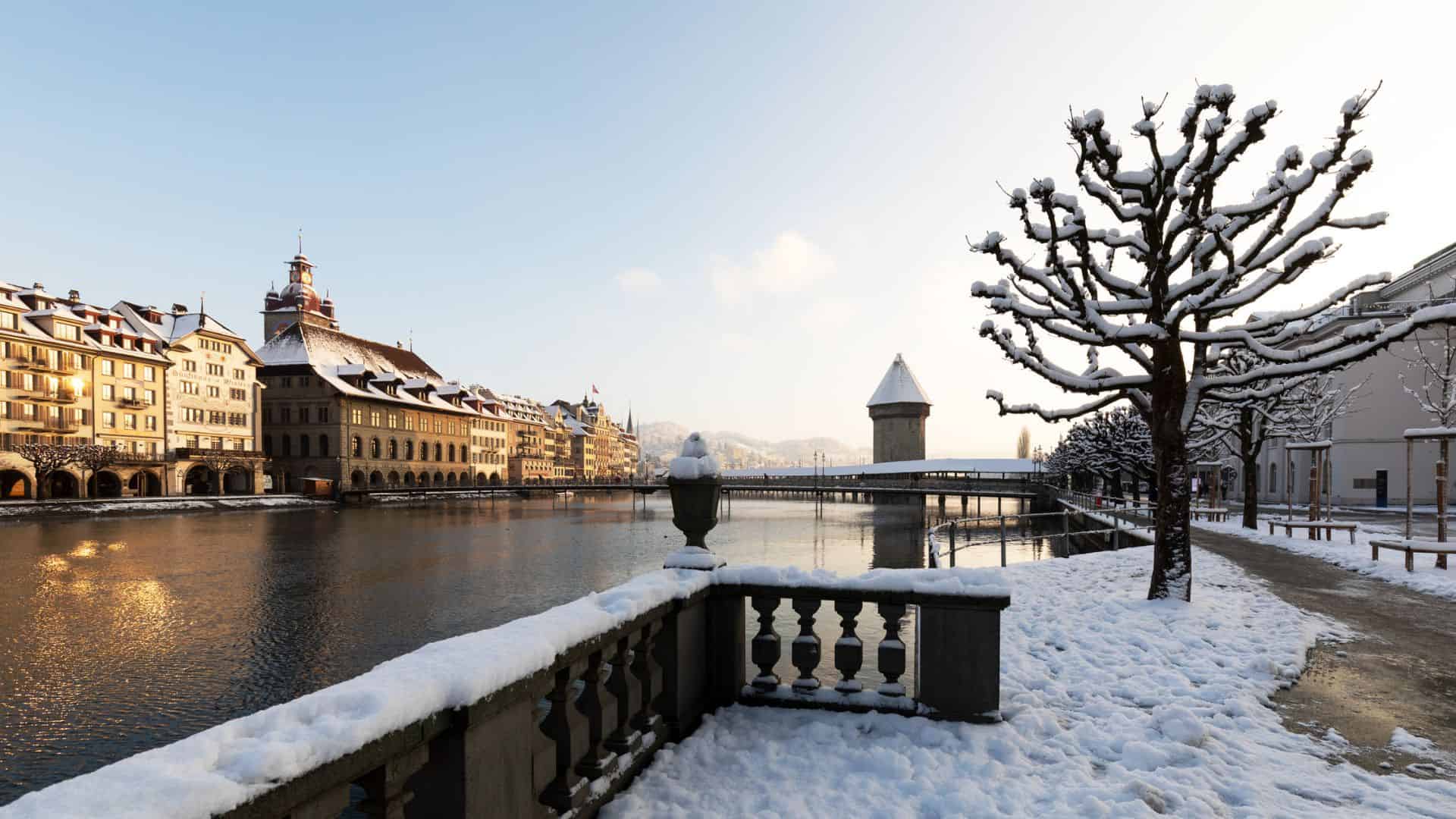 Snow-covered riverside promenade with bare trees, historic buildings, and a stone tower in winter sunlight.