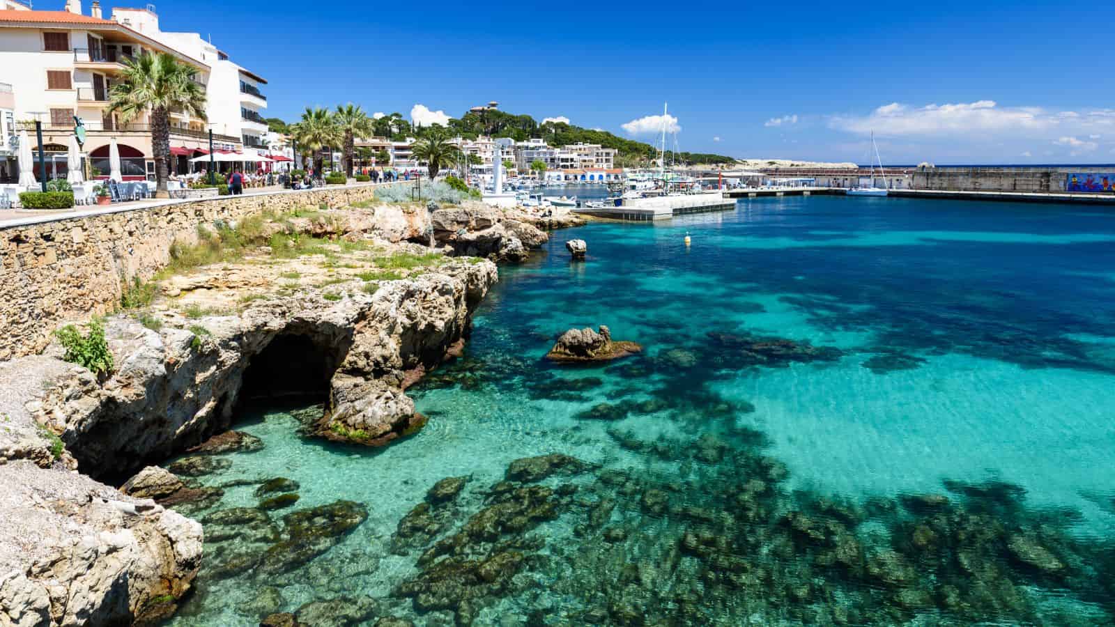 Clear turquoise water by a rocky shore with buildings and palm trees along a sunny seaside promenade.