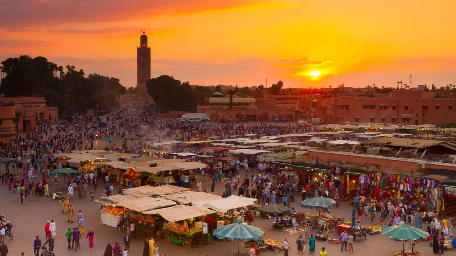 Crowded marketplace in Marrakech at sunset, with vibrant stalls and a tall tower in the background.