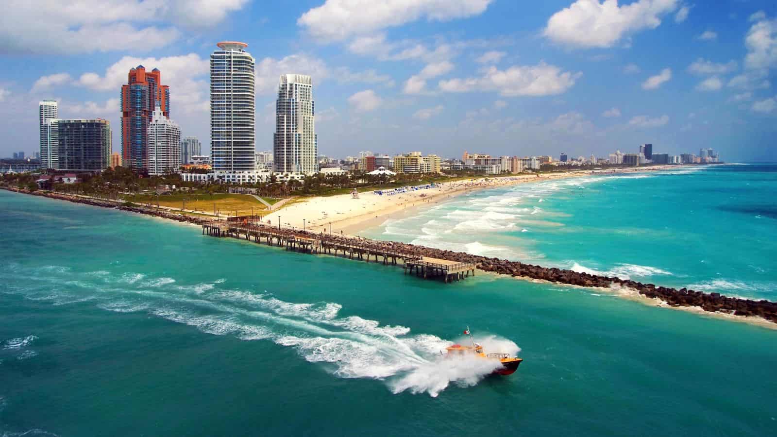 A speedboat moves near a pier by a sandy beach with tall buildings and blue ocean under a partly cloudy sky.