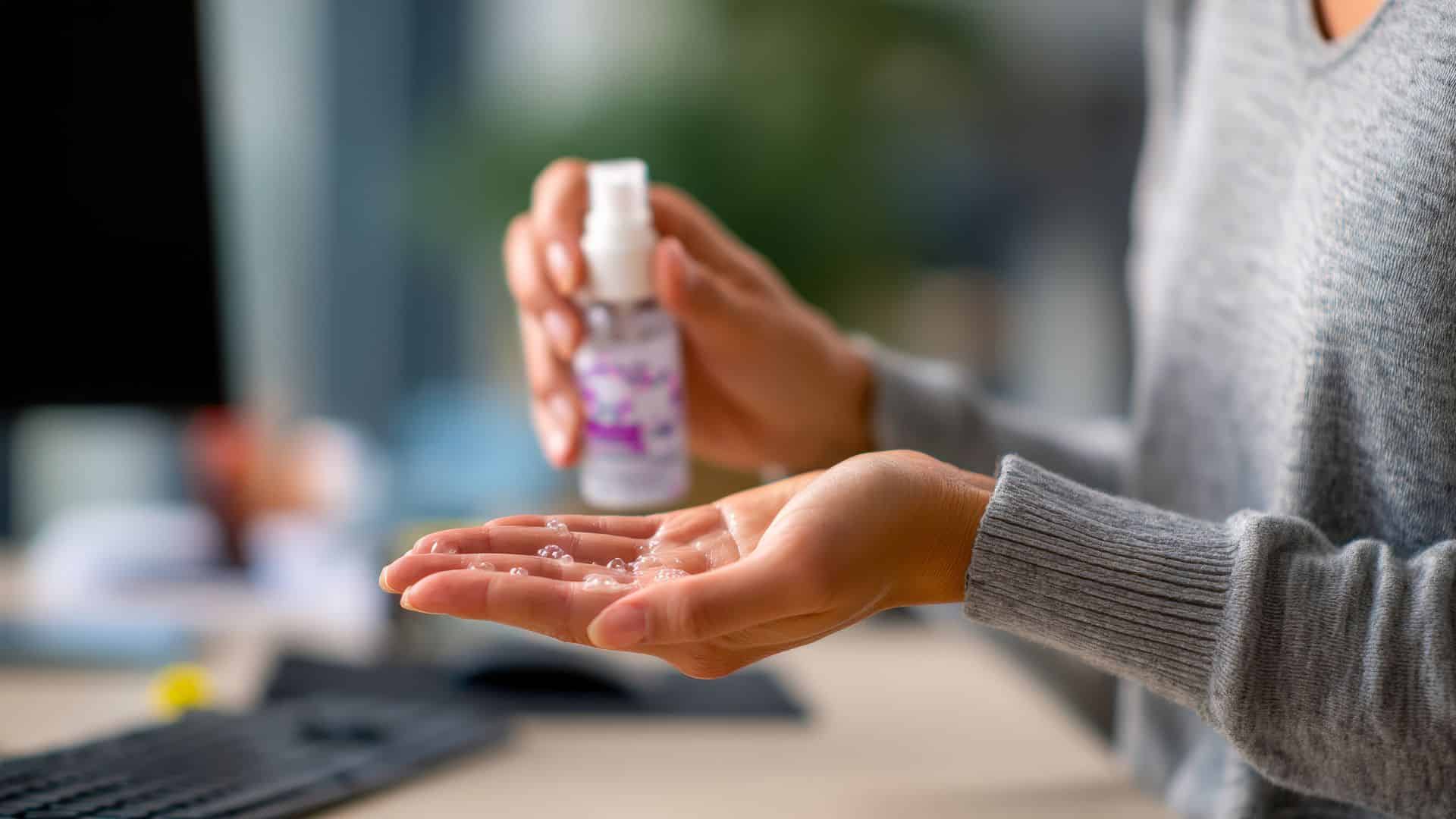 Person applying hand sanitizer from a small bottle, sitting at a desk with a keyboard in the background.